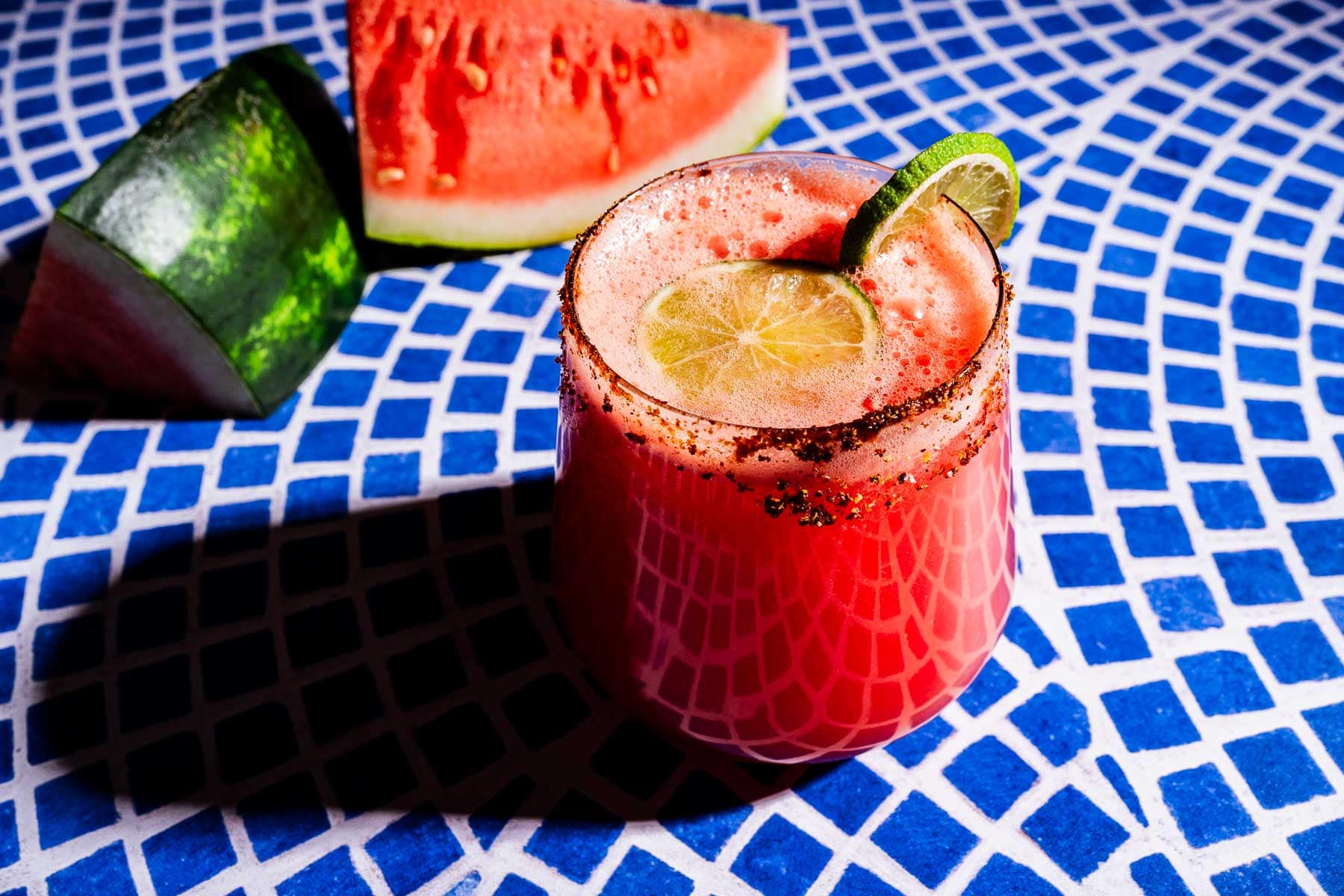 A glass of red watermelon agua fresca with a chili-rimmed glass and lime slices, set on a blue patterned table with watermelon wedges in the background.
