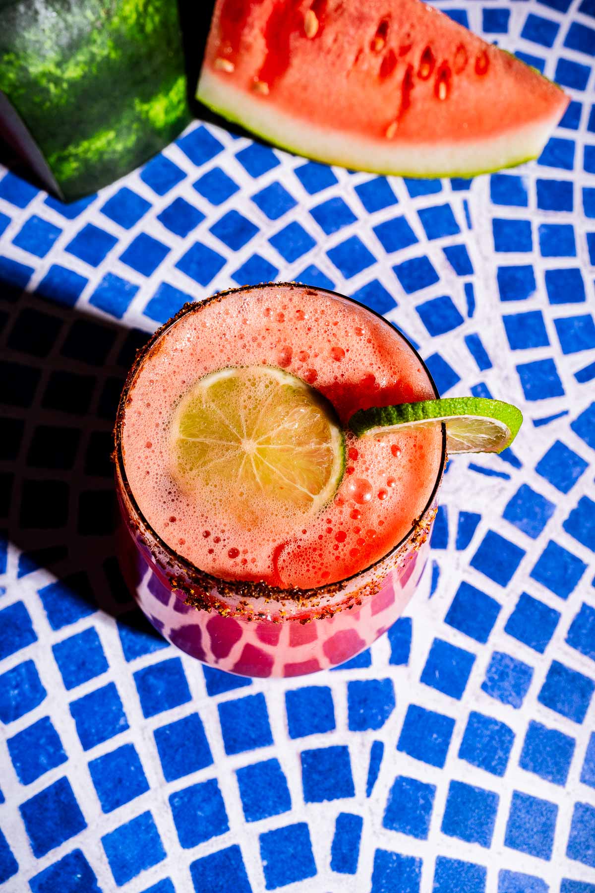 A vibrant watermelon agua fresca cocktail with a lime slice garnish sits on a blue and white mosaic table; a wedge of watermelon is in the background.