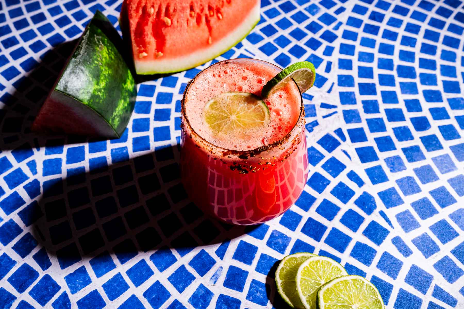 A glass of red watermelon agua fresca garnished with lime sits on a blue patterned surface, with watermelon slices and lime rounds nearby.