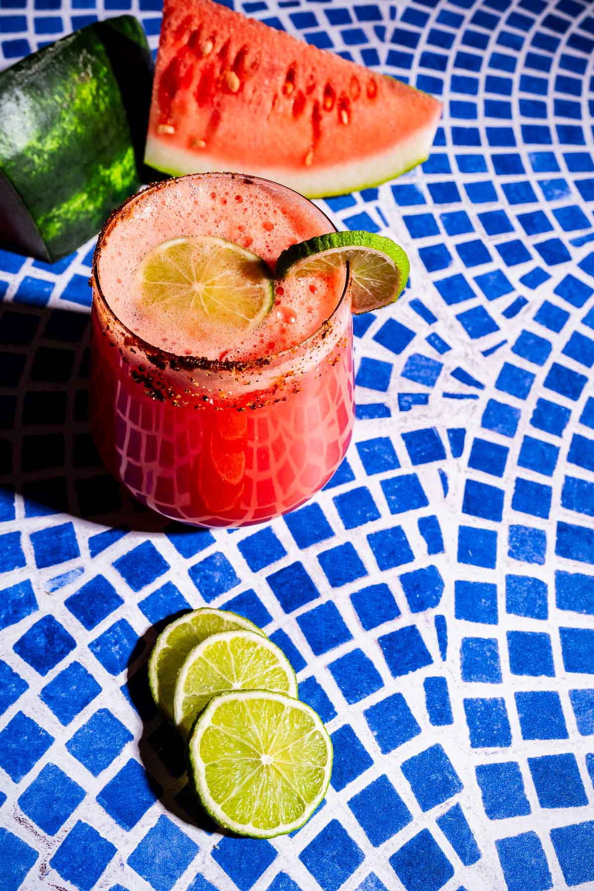 A glass of watermelon agua fresca garnished with a lime wheel sits on a blue mosaic table, with lime slices and watermelon pieces nearby.