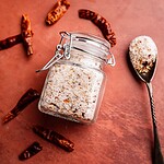 A glass jar filled with spicy salt sits on a brown surface, surrounded by dried red chili peppers, with a spoonful of the chili salt nearby.