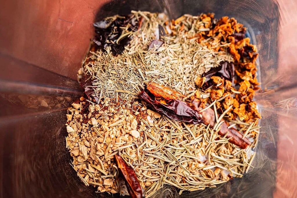 A close-up of various dried herbs and spices, including chili peppers, rosemary, chopped ginger, and spicy salt, inside a transparent container.