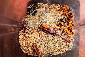 A close-up of various dried herbs and spices, including chili peppers, rosemary, chopped ginger, and spicy salt, inside a transparent container.