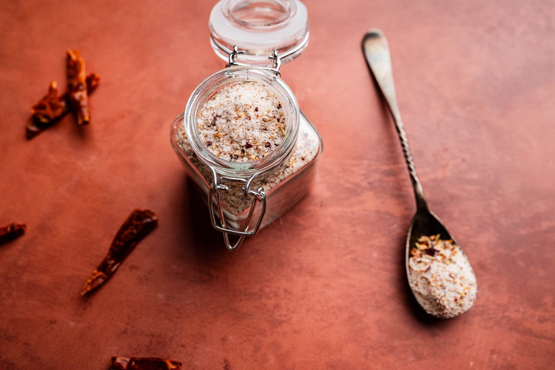 A glass jar of spicy salt sits open on a reddish surface, with a spoonful of the mixture nearby and dried chili peppers scattered around.