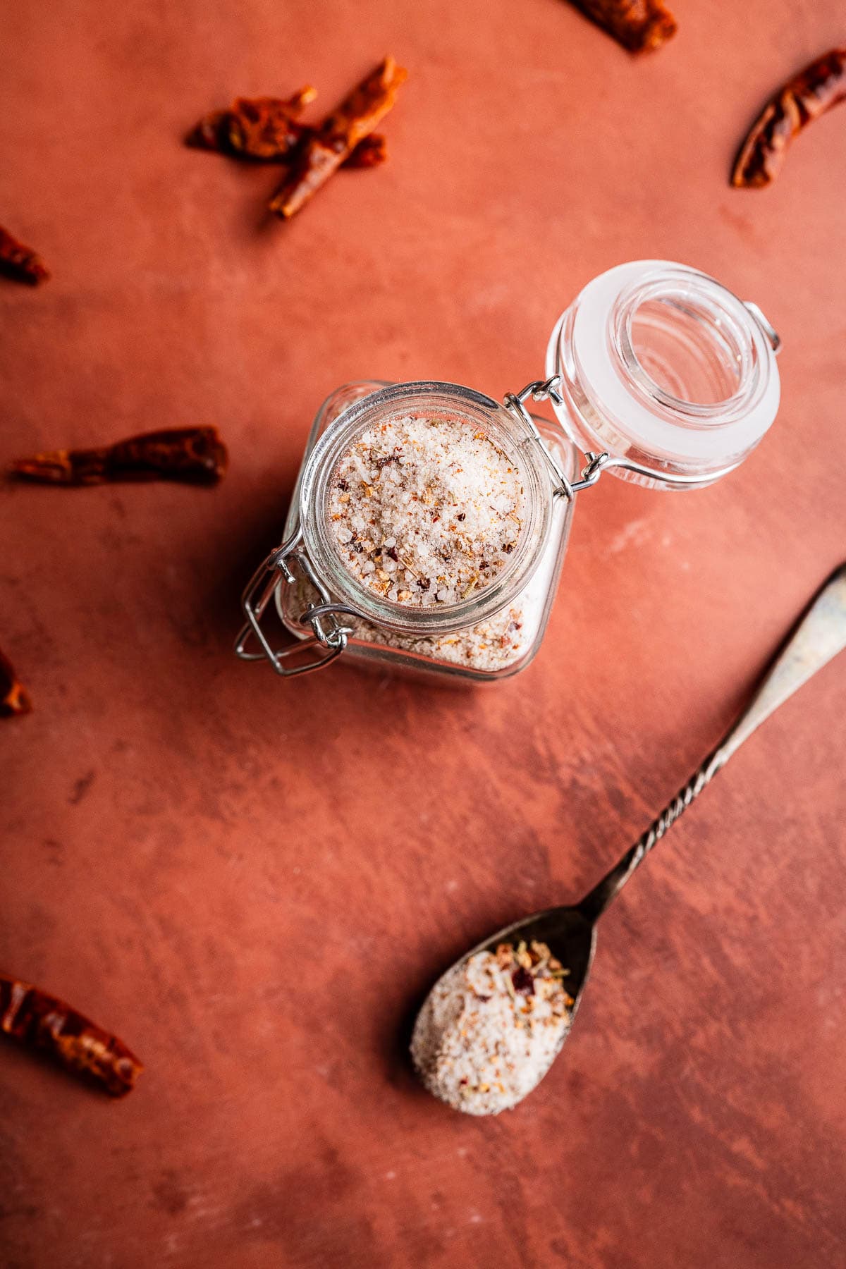 A glass jar filled with spicy salt sits open on a brown surface, surrounded by dried chili pieces, with a spoonful of the salt nearby.