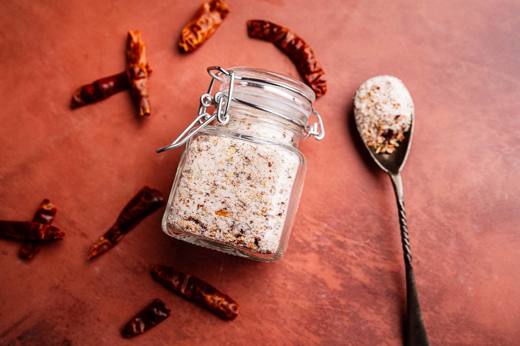 A glass jar of spicy salt blend sits on a brown surface next to a spoonful of the mix and several dried red chili peppers scattered around.