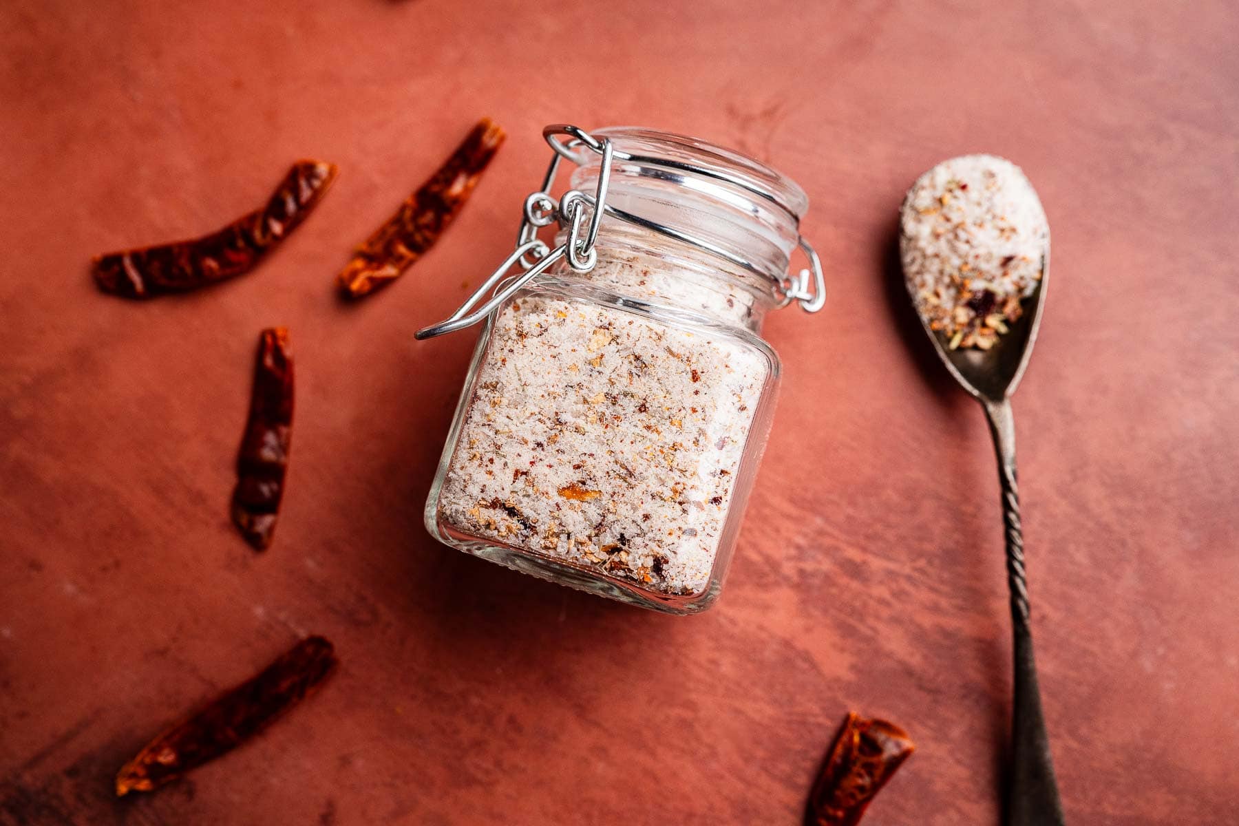 A glass jar filled with spicy salt sits on a brown surface, surrounded by dried chili peppers, with a spoonful of the spicy salt nearby.