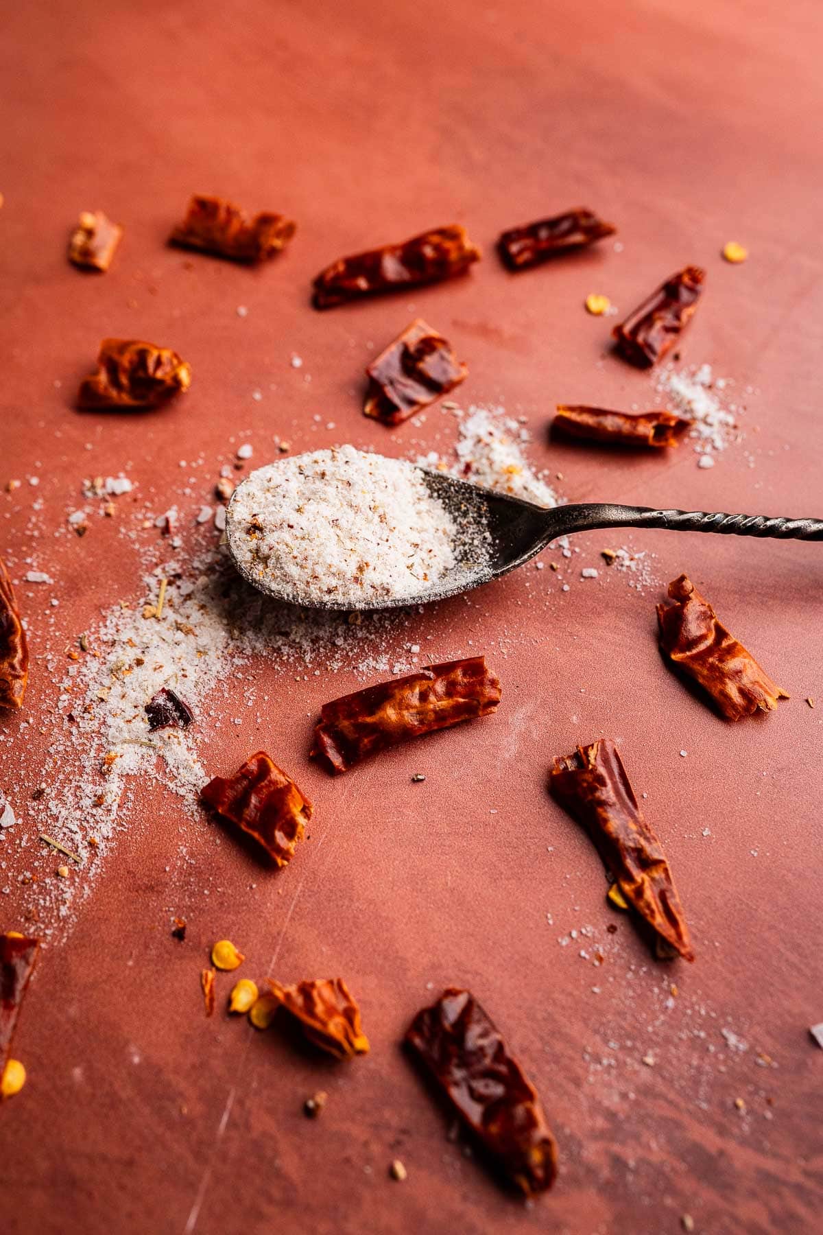 A spoon filled with spicy salt rests on a reddish surface, surrounded by scattered dried chili pieces and salt.
