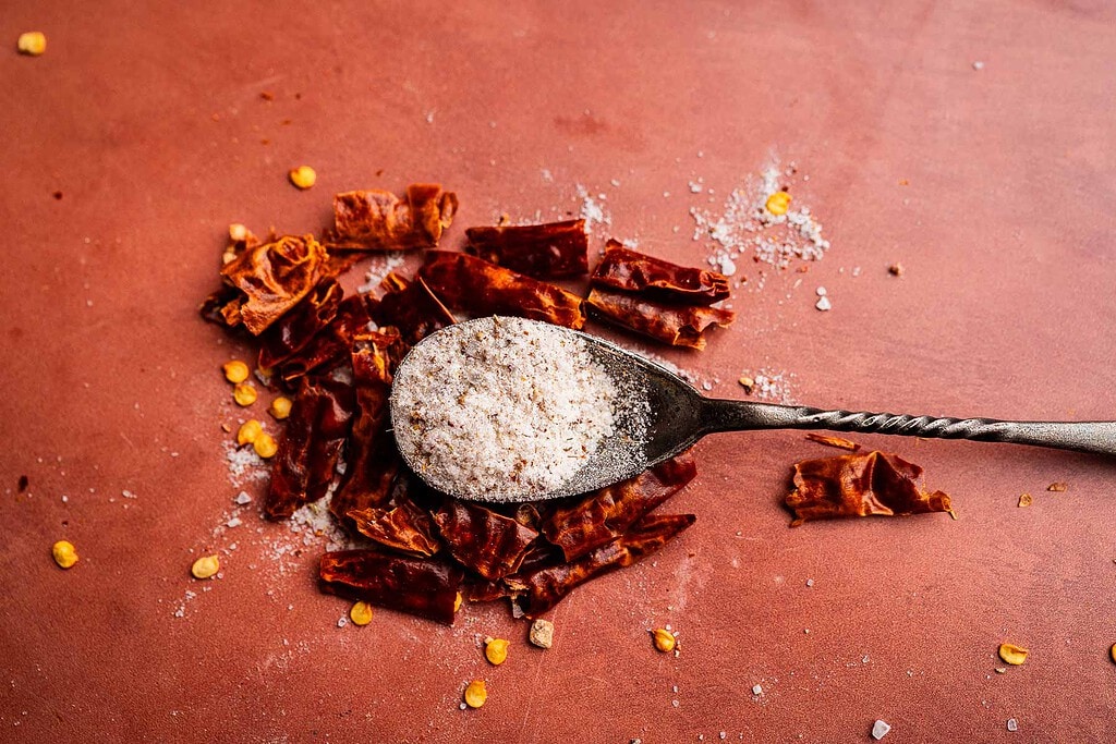 A metal spoon with spicy salt rests on top of chopped dried red chili peppers, with some salt and seeds scattered on a brown surface.