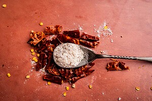 A metal spoon with spicy salt rests on top of chopped dried red chili peppers, with some salt and seeds scattered on a brown surface.