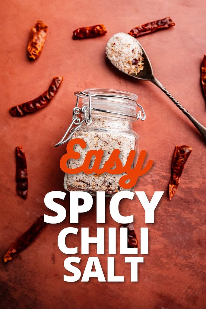 A jar of spicy salt with a spoonful beside it, surrounded by dried chili peppers on a reddish surface. Text reads "Easy Spicy Chili Salt.