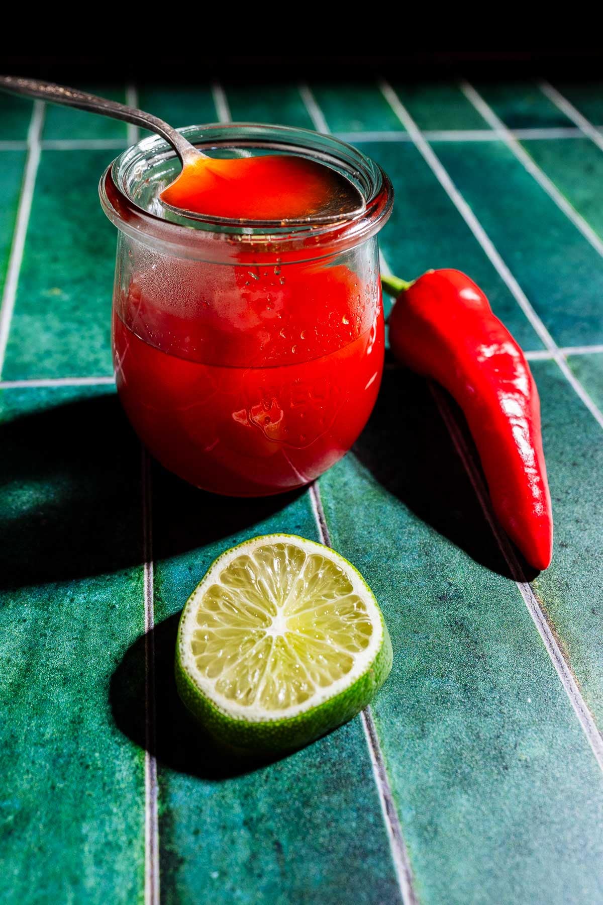 A glass jar of spicy watermelon syrup with a spoon inside sits on a green tiled surface next to a red chili pepper and a halved lime.
