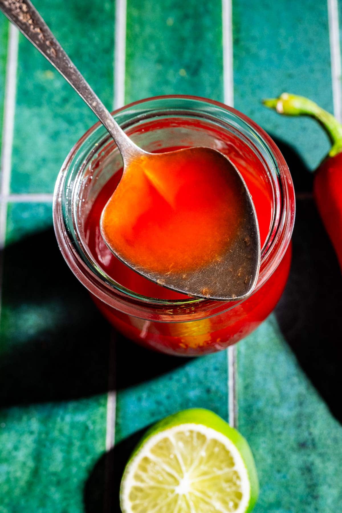 A spoon holds red hot sauce above a glass jar, with a chili pepper and half a lime on a green tiled surface nearby—perfect inspiration for making your own spicy watermelon syrup.