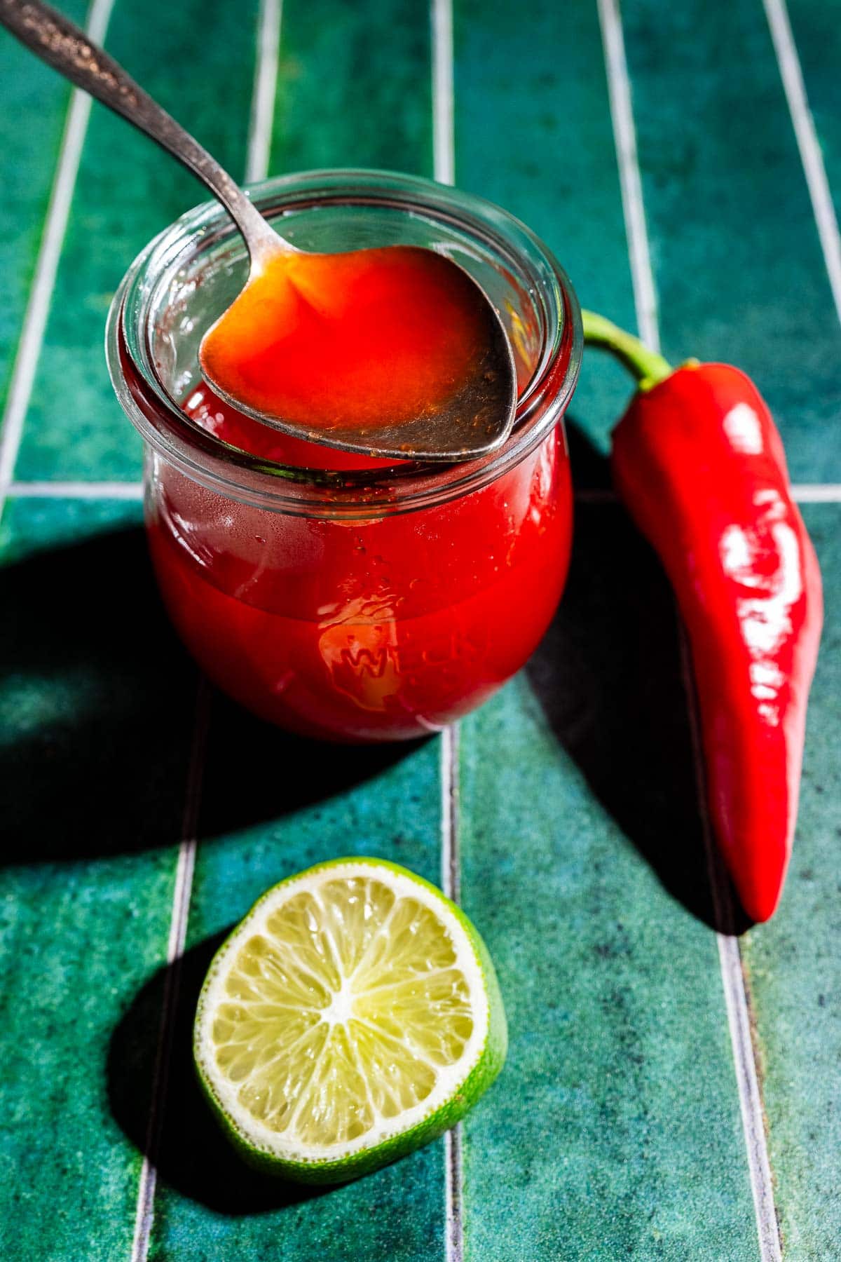 A glass jar of red sauce with a spoon, next to a red chili pepper and a halved lime on a green tiled surface, evokes the vibrant look of spicy watermelon syrup.