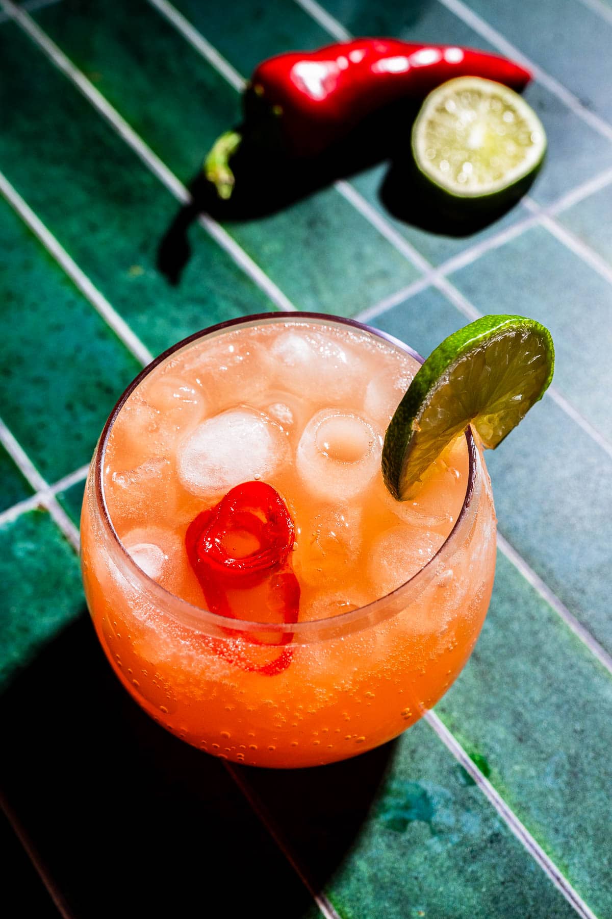 A round glass of spicy watermelon mocktail with ice, garnished with a lime wheel and red chili, sits on a green tiled surface. A halved lime and whole chili are in the background.