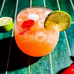 A round glass filled with a spicy watermelon mocktail, orange fizzy drink, ice cubes, a lime wheel on the rim, and a red garnish, placed on a green striped surface with a cut lime nearby.