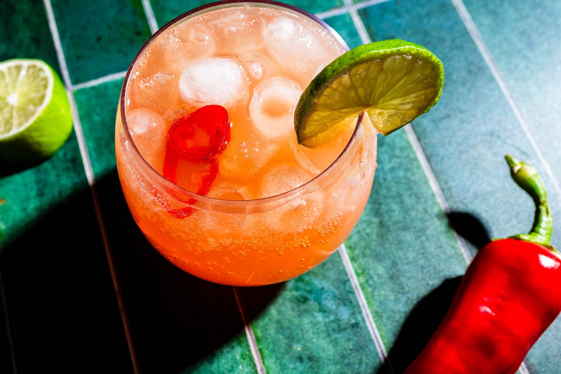 A glass of iced orange cocktail garnished with a lime slice and red chili peppers sits on a green tiled surface beside a halved lime and a whole red chili pepper—a refreshing twist on the spicy watermelon mocktail.