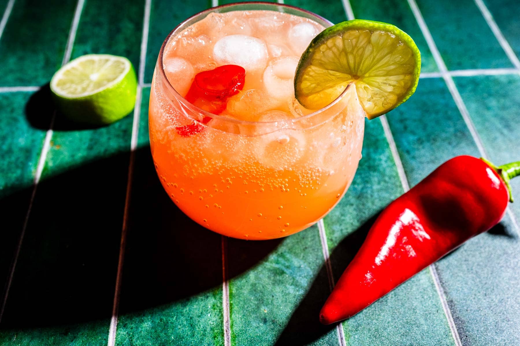 A glass of spicy watermelon mocktail with ice, a lime wheel garnish, and a red chili pepper inside, placed on a green tiled surface with a chili pepper and a lime half nearby.