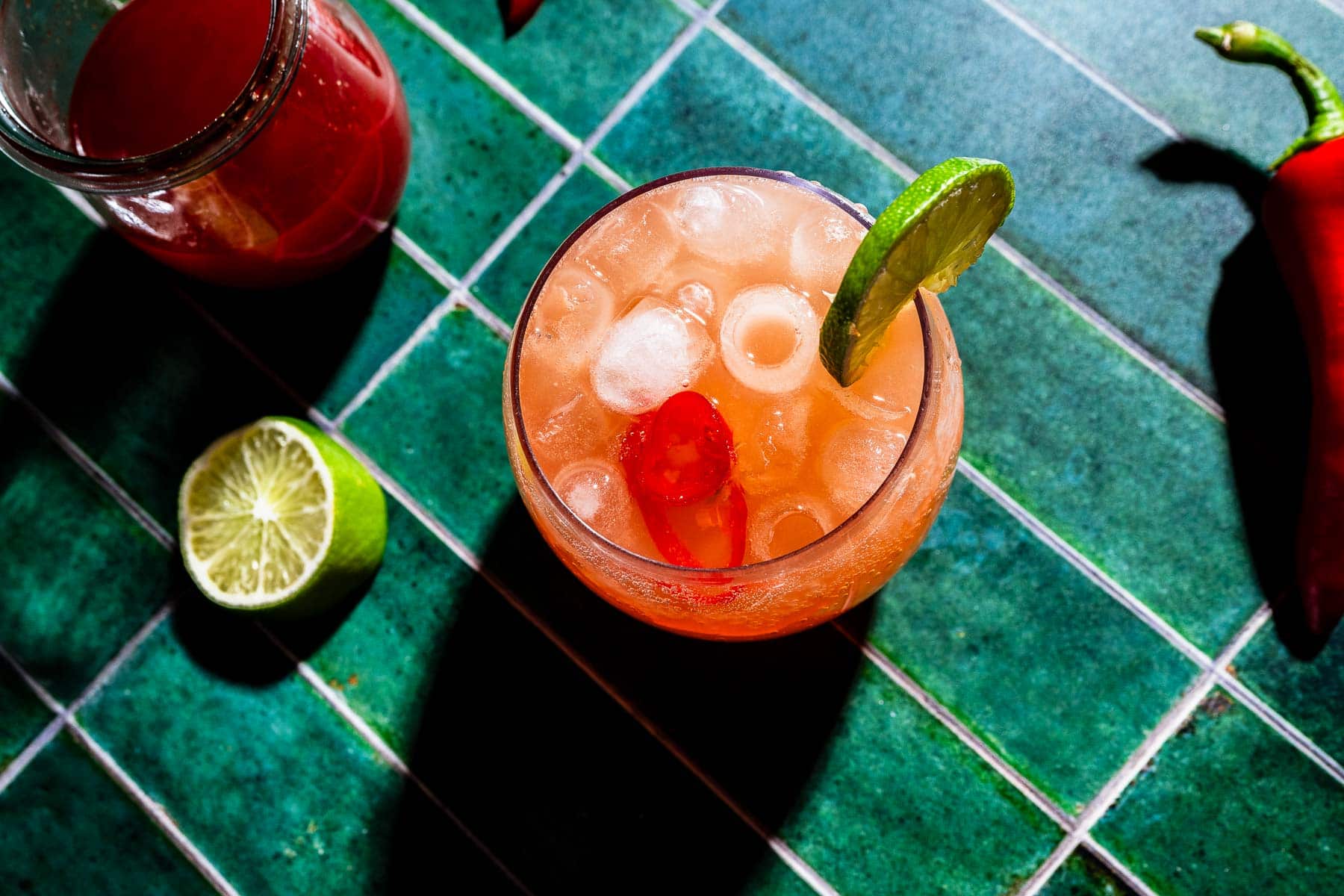 A glass of orange-pink spicy watermelon mocktail, garnished with a lime wedge and cherry, sits on a green tiled surface beside a sliced lime, drink jar, and chili pepper.