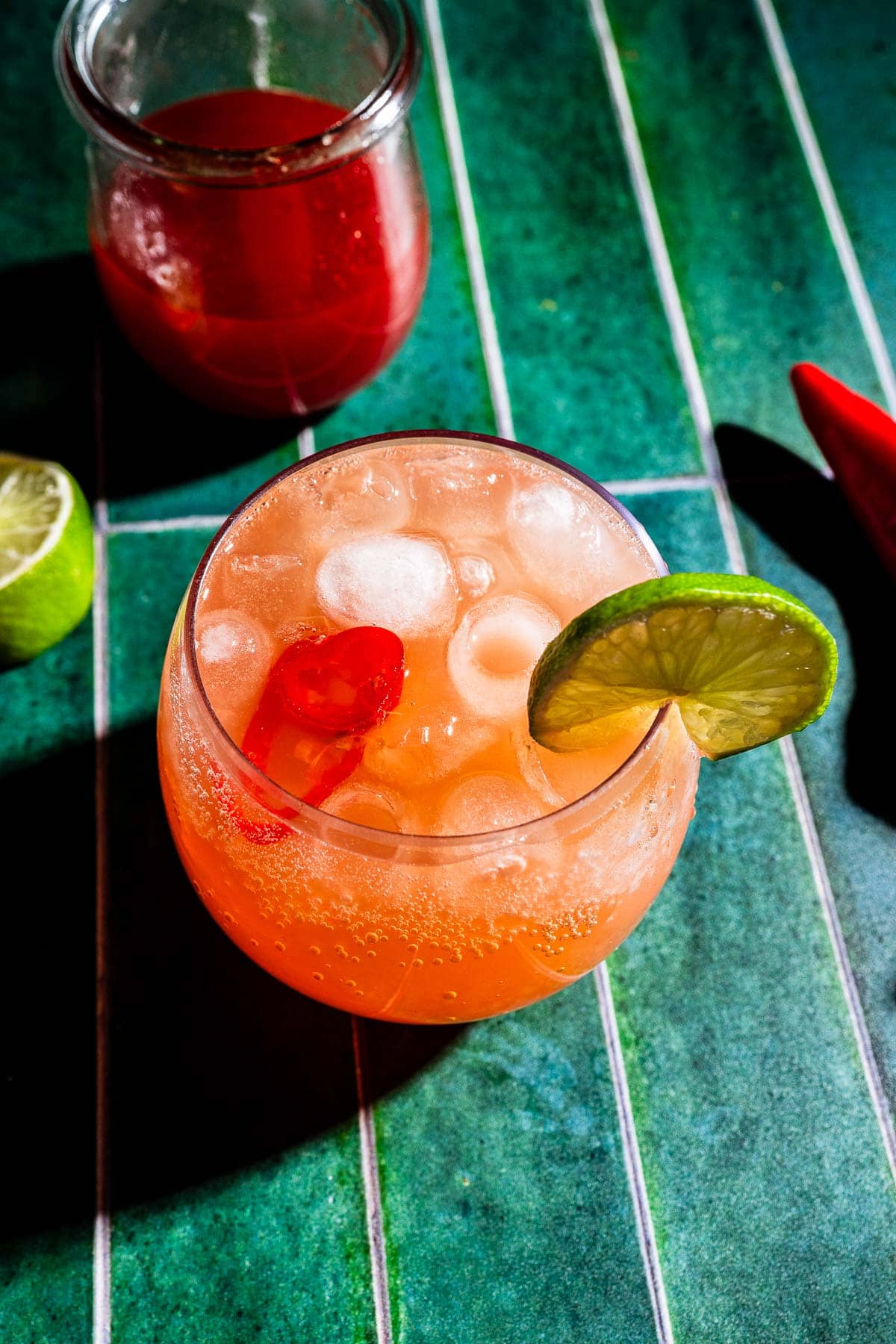 A glass of spicy watermelon mocktail garnished with a lime slice and red chili, with a jar of red liquid and half a lime in the background on a green tiled surface.