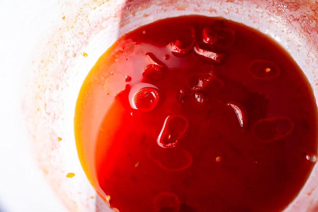 A close-up of a white bowl containing red sauce with visible sliced red chilies floating on the surface, hinting at the bold flavor of spicy watermelon syrup.