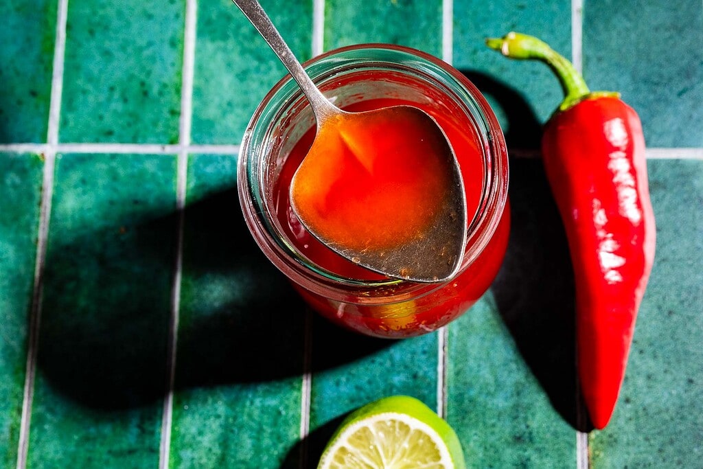 A jar of spicy watermelon syrup with a spoon inside sits beside a whole red chili pepper and a halved lime on a green tiled surface.
