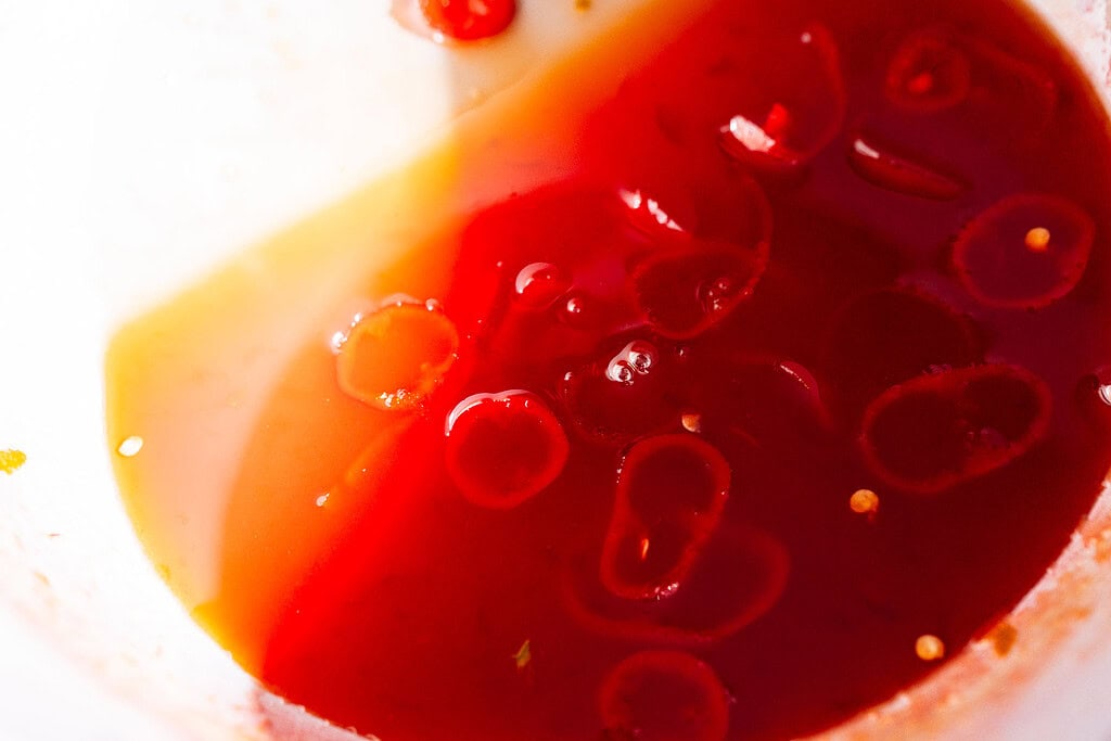 Close-up of a red liquid with oil and chili slices floating on the surface, likely a spicy watermelon syrup or dressing in a bowl.