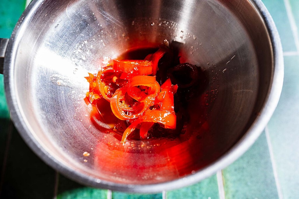 Sliced red peppers in a small amount of spicy watermelon syrup inside a stainless steel mixing bowl on a green tiled surface.