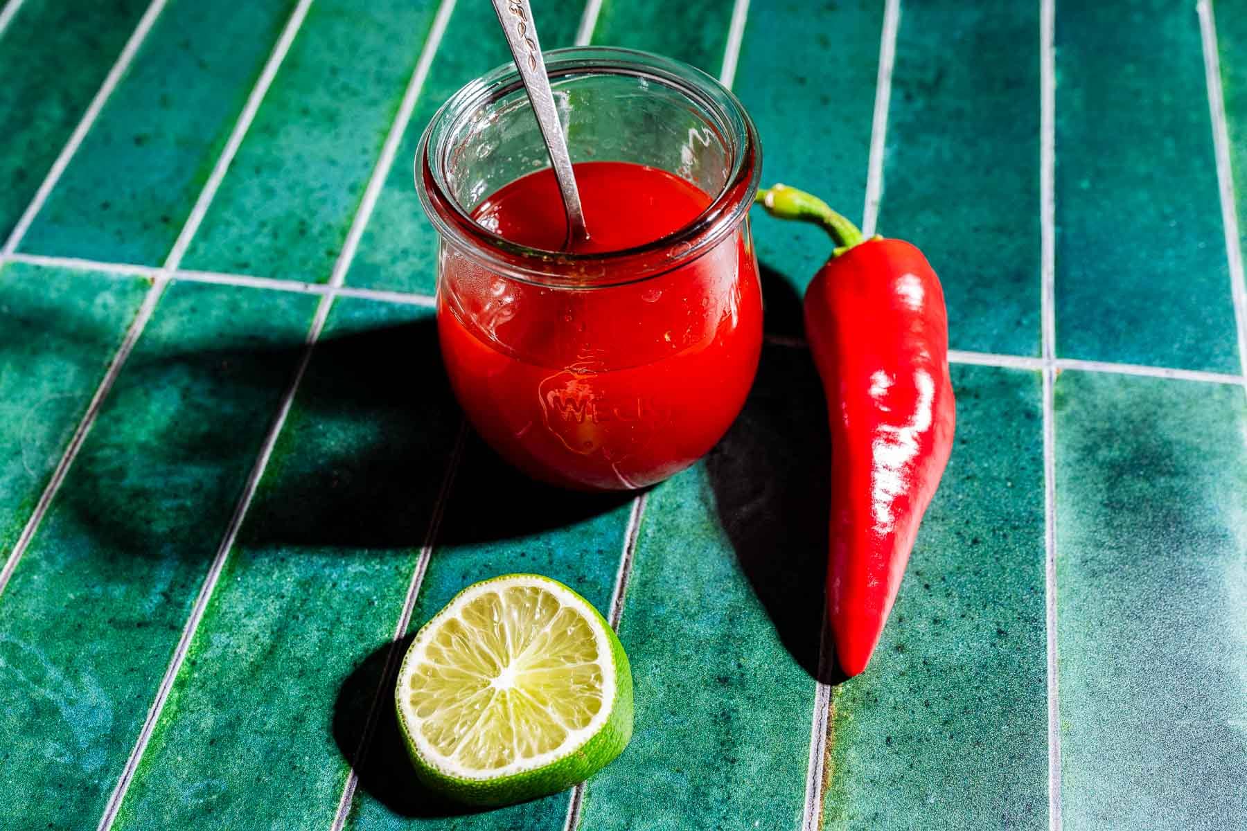 A small jar of red sauce with a spoon, a red chili pepper, and a halved lime on a green tiled surface—perfect for drizzling spicy watermelon syrup on your favorite dishes.