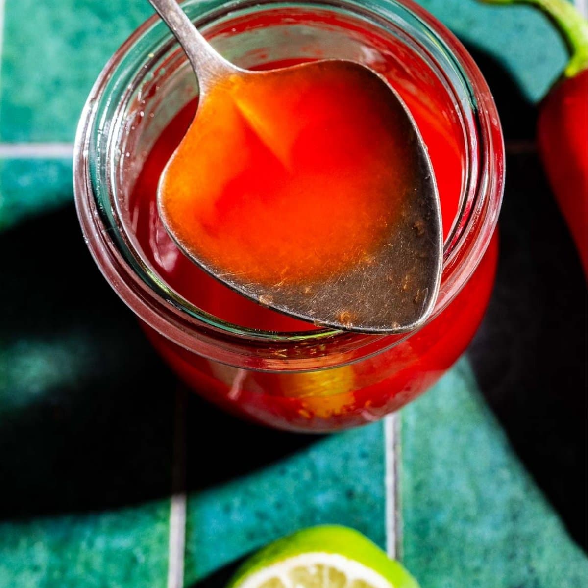 A spoon rests on a jar filled with red liquid, likely spicy watermelon syrup, placed on a green tiled surface with a lime slice nearby.