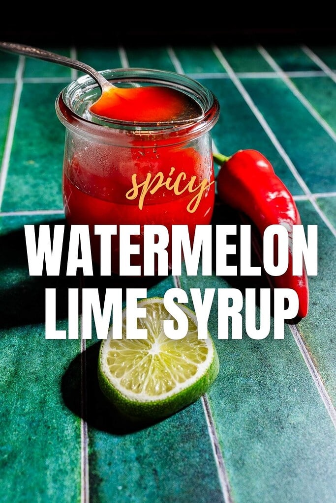 A jar of spicy watermelon syrup with a spoon, a red chili pepper, and a halved lime sits on a green tiled surface, highlighting the tangy blend of watermelon and lime with a kick of heat.
