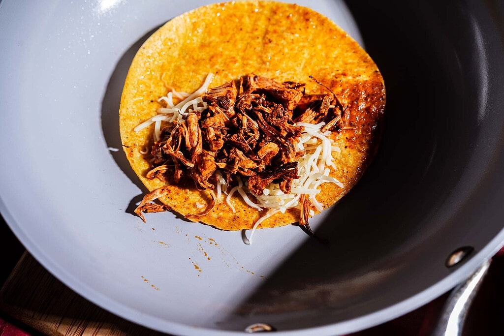 A corn tortilla with shredded cheese and seasoned jackfruit birria filling is in a skillet, ready to be cooked.