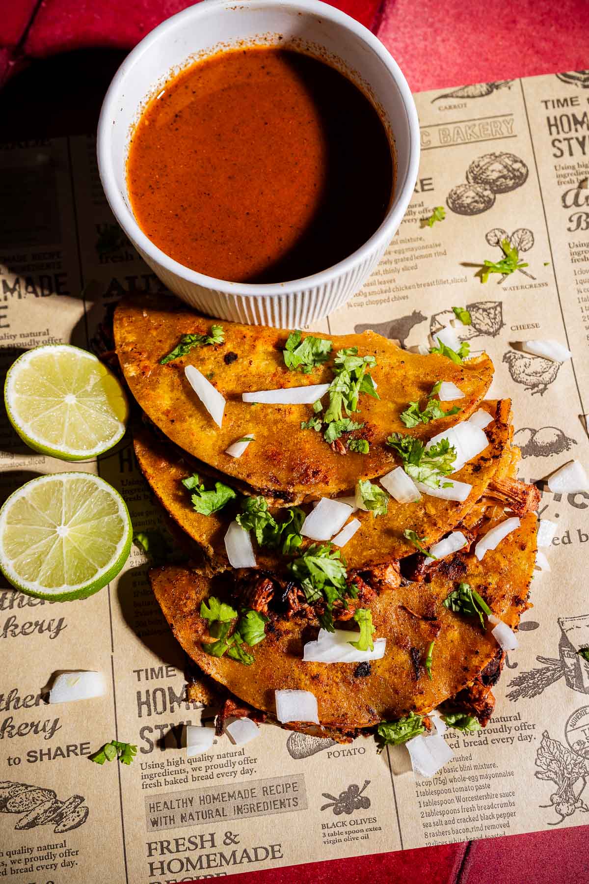 Three jackfruit birria tacos topped with chopped onions and cilantro, served with lime halves and a cup of consommé on a printed paper background.
