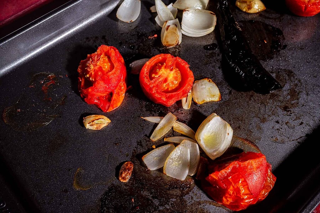 Roasted tomatoes, onions, garlic, and a dark pepper rest on a baking tray with visible oil and charred spots, perfectly prepped for rich jackfruit birria.