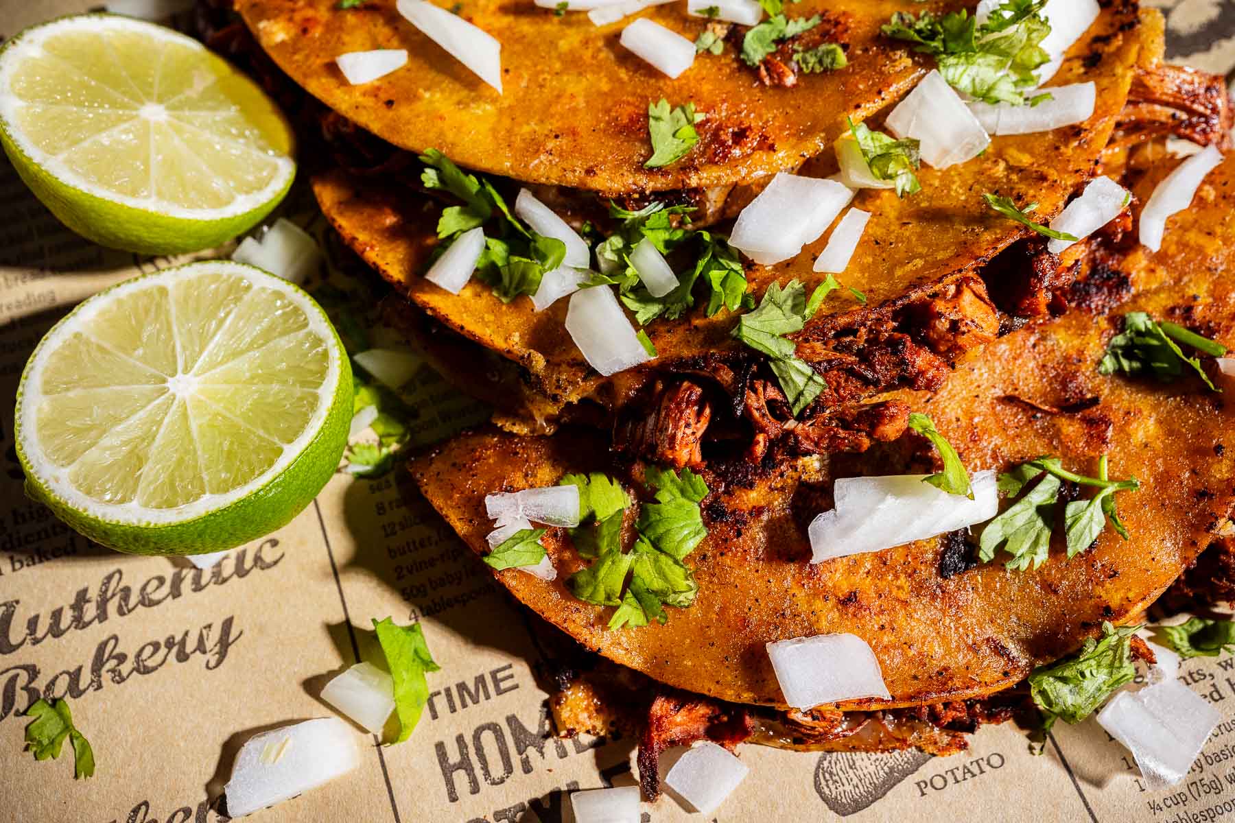 Three tacos filled with savory jackfruit birria, topped with chopped onions and cilantro, served on newspaper with two lime halves on the side.