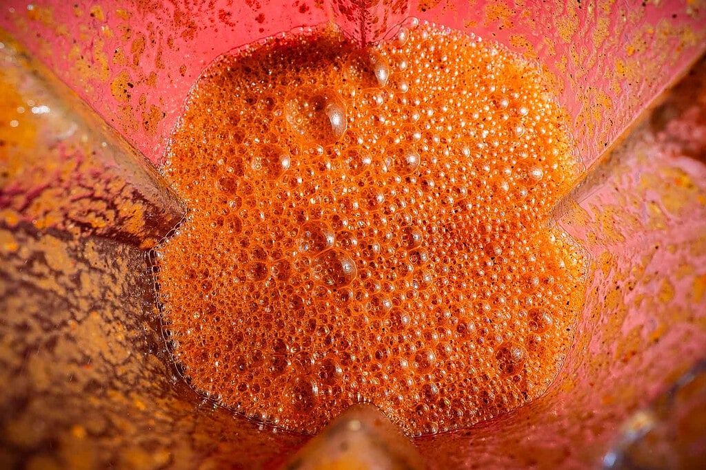 Close-up view of a red-orange, frothy jackfruit birria liquid inside a blender, with visible bubbles and residue on the blender walls.