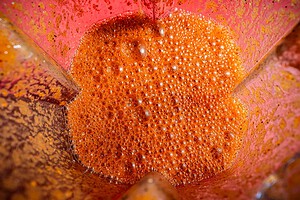 Close-up view of a red-orange, frothy jackfruit birria liquid inside a blender, with visible bubbles and residue on the blender walls.