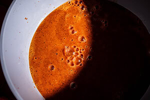 A close-up of jackfruit birria, its reddish-brown liquid with bubbles on the surface, partially shaded inside a white bowl.
