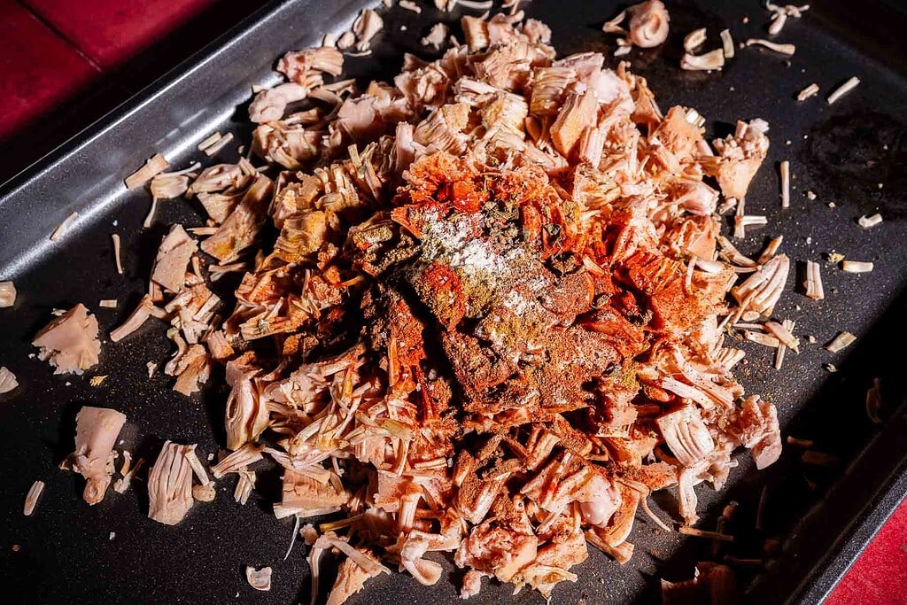 Shredded jackfruit, perfect for jackfruit birria, is topped with various spices on a dark baking tray, ready for cooking.
