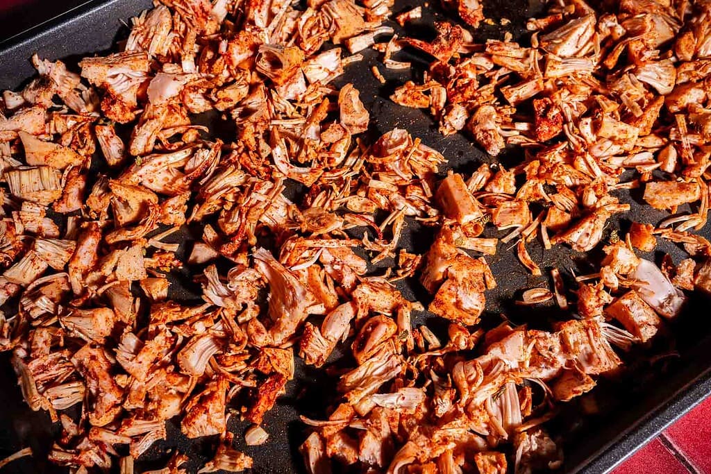 Seasoned, shredded jackfruit for jackfruit birria spread out on a dark baking tray, ready for roasting.