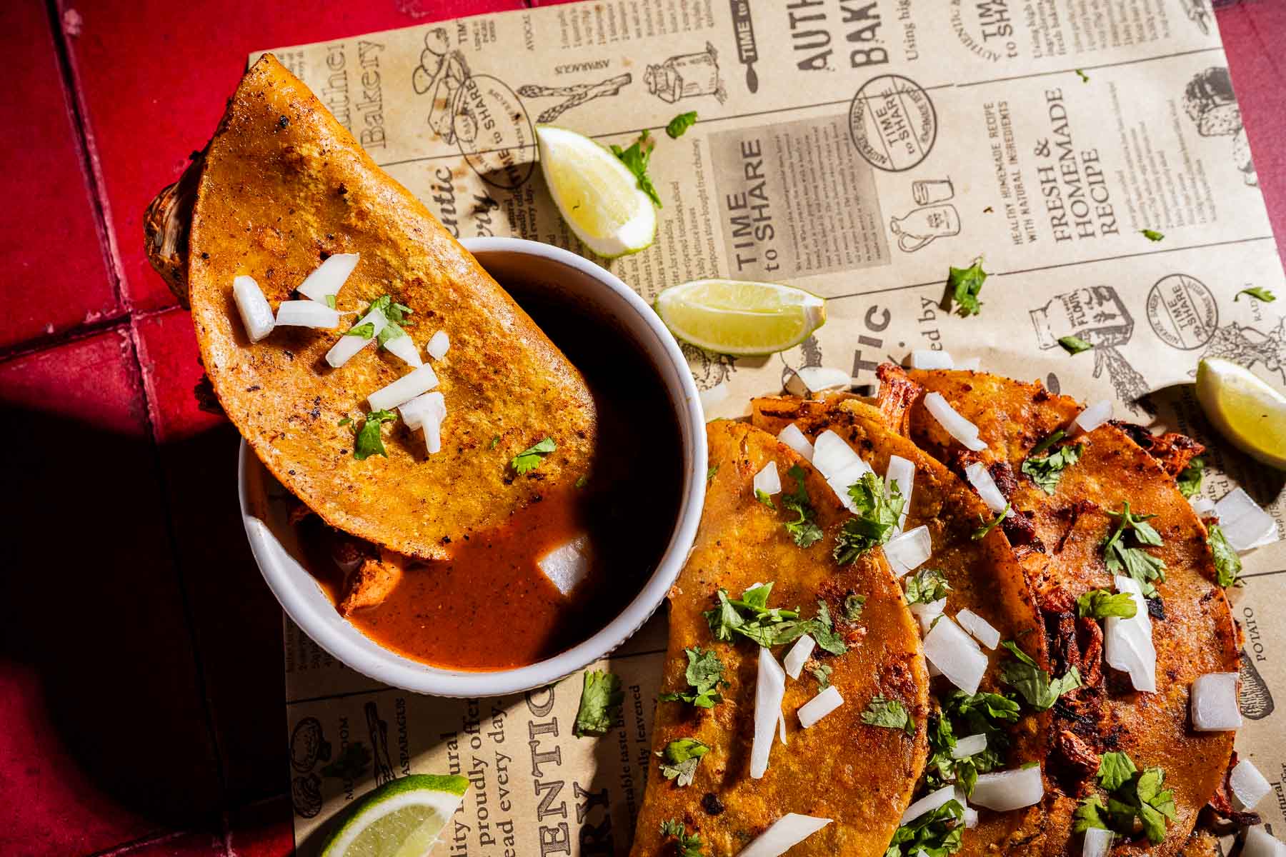 Three jackfruit birria tacos topped with chopped onions and cilantro, served with lime wedges and a bowl of consommé on newspaper-style parchment paper.