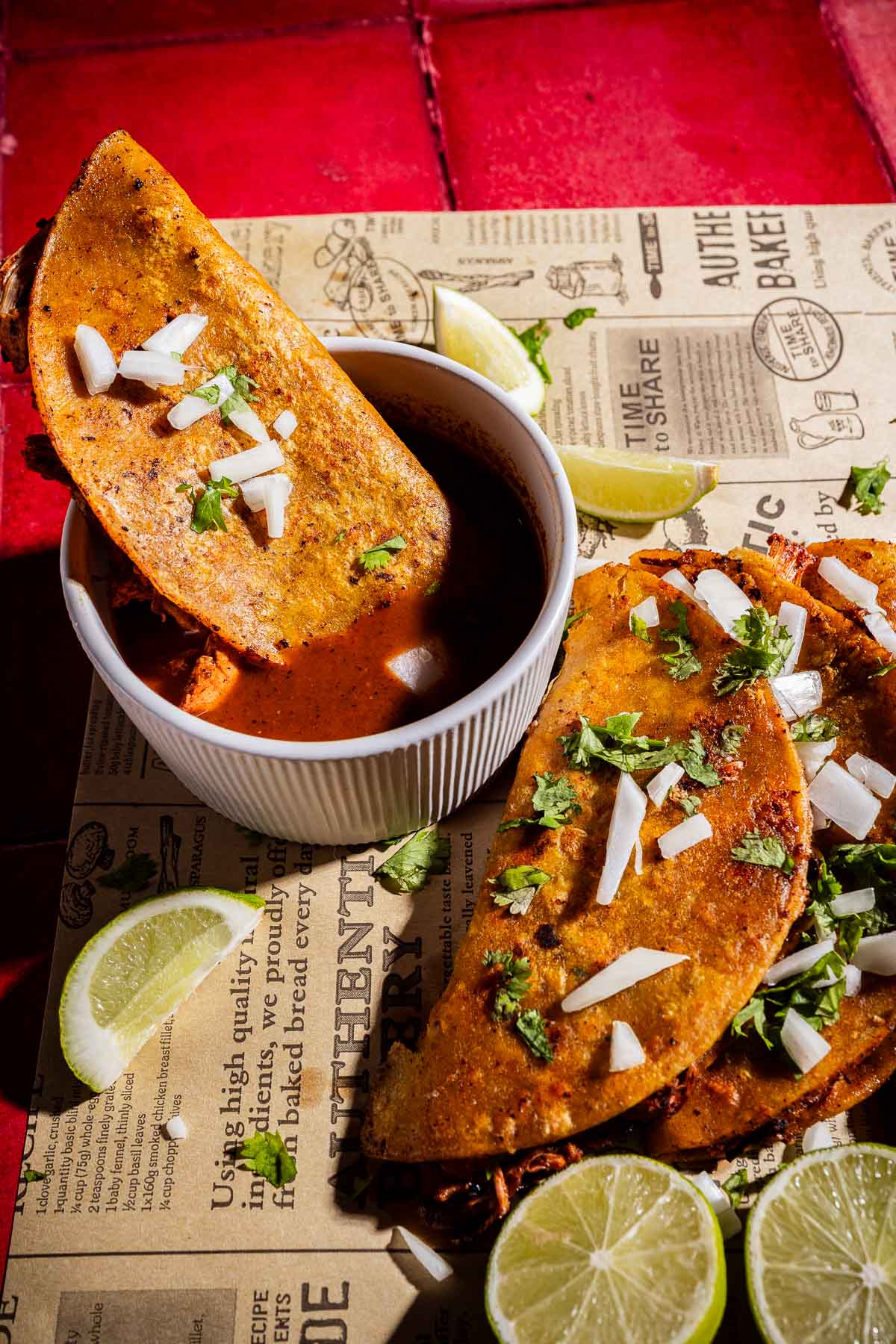 Three jackfruit birria tacos topped with chopped onions and cilantro are served with lime wedges and a bowl of dipping consommé on patterned paper.