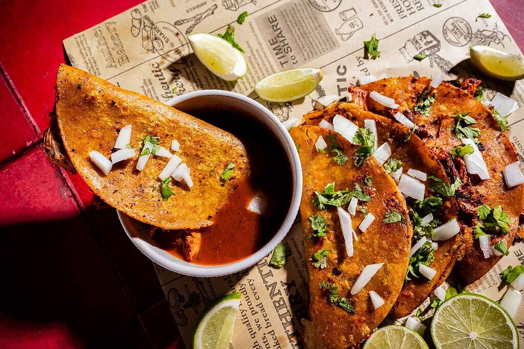 Three jackfruit birria tacos topped with chopped onions and cilantro are served with lime wedges and a bowl of consommé on a sheet of decorative paper.