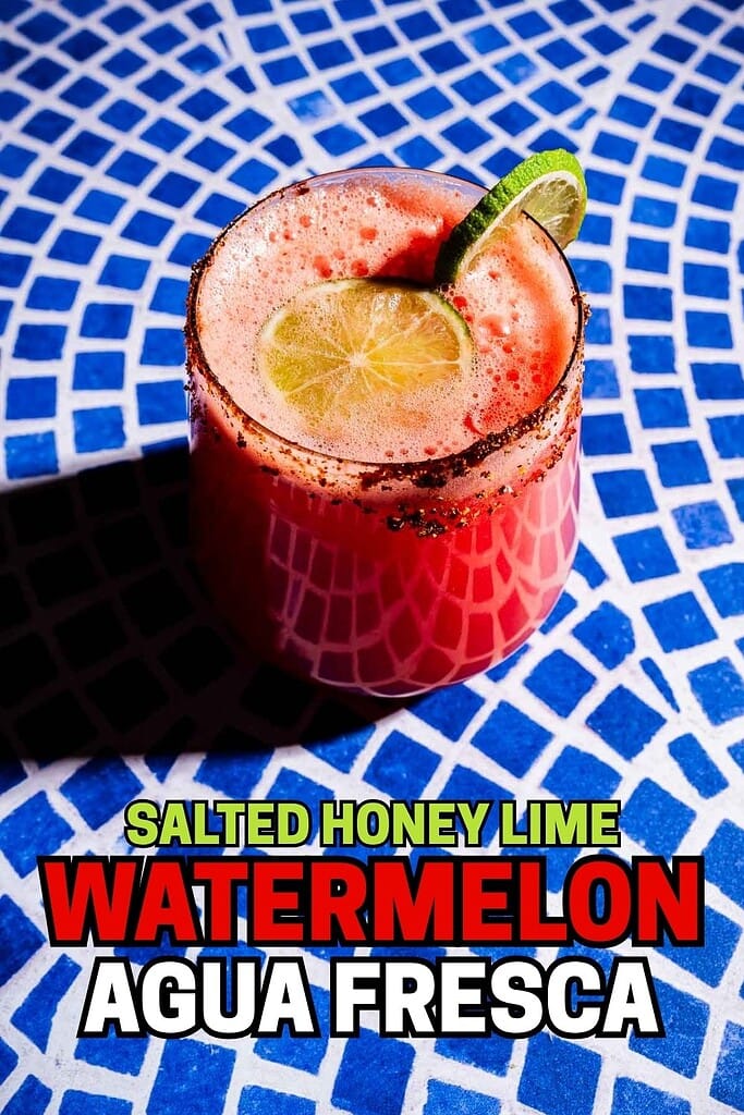 A refreshing glass of salted honey lime watermelon agua fresca with a lime slice on the rim sits on a blue and white tiled surface.