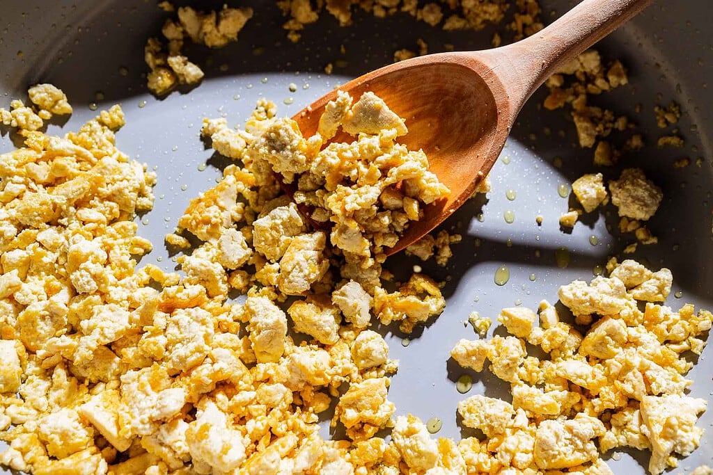 Scrambled tofu, reminiscent of sofritas, is being cooked in a pan with a wooden spoon, with some oil visible.
