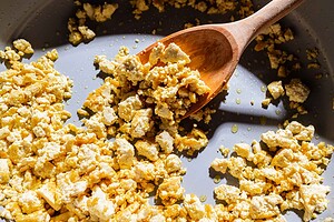 Scrambled tofu, reminiscent of sofritas, is being cooked in a pan with a wooden spoon, with some oil visible.