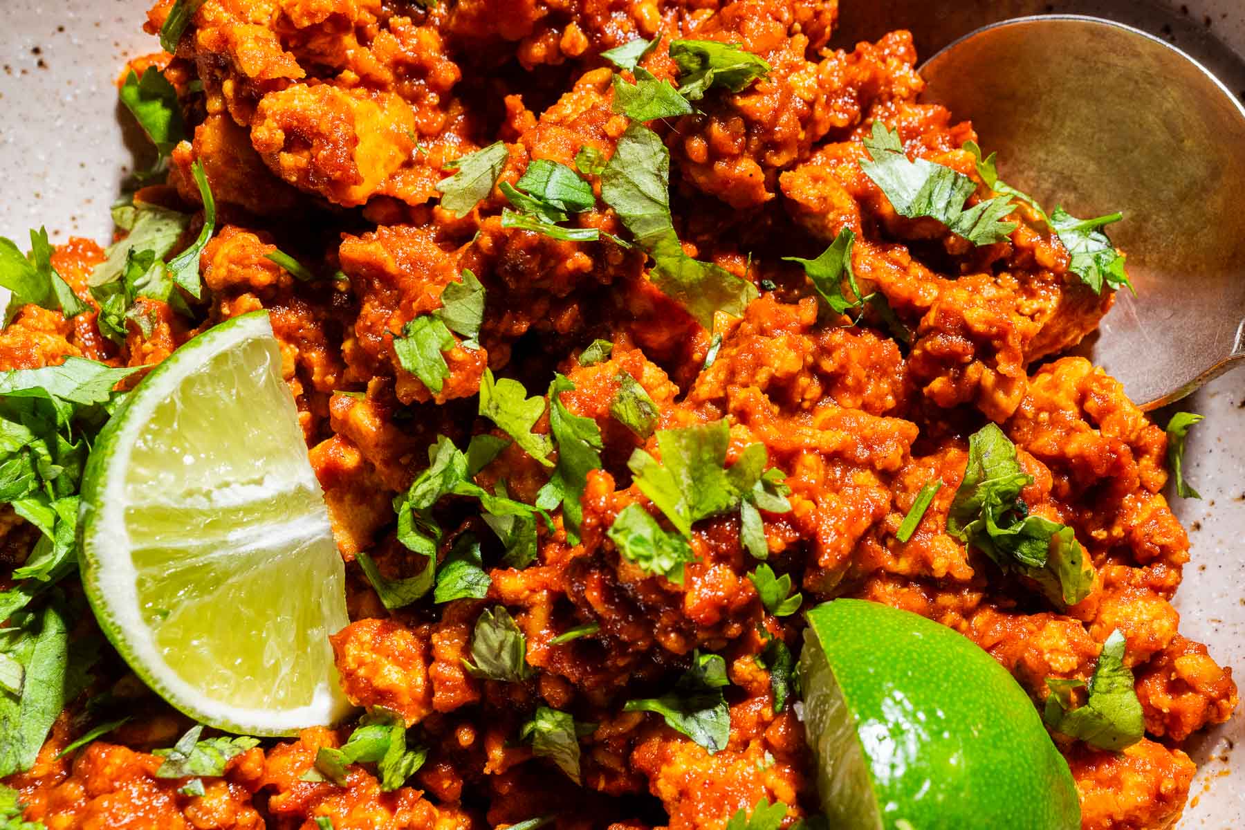 Close-up of a spicy sofritas dish topped with chopped cilantro, served with lime wedges and a metal spoon.