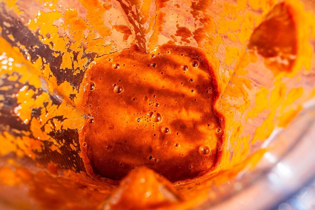 A close-up view of thick red sofritas sauce or paste with bubbles inside a blender jar, showing remnants on the sides.