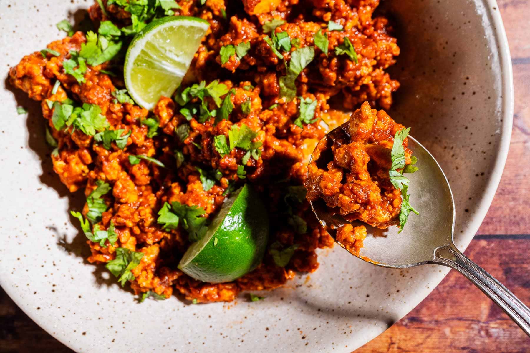 A bowl of seasoned sofritas topped with chopped cilantro and lime wedges, with a spoon lifting a portion.