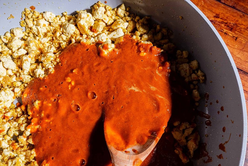 Crumbled tofu in a pan with savory sofritas red sauce is being stirred with a wooden spoon on a wooden surface.